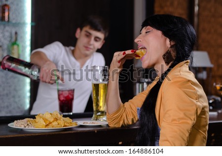 Young woman enjoying a snack at the bar sitting eating chips with a glass of beer close by as the bartender works preparing drinks in the background
