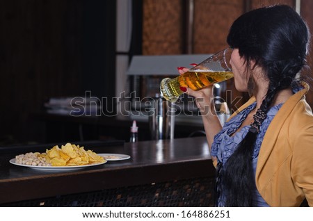 View from behind of a young woman gulping down a pint beer at the bar during Happy Hour with a plate of snacks on the counter in front of her