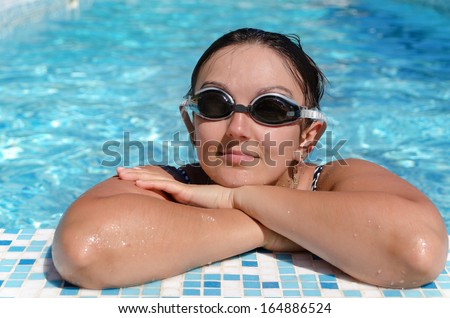 Woman relaxing at the side of a swimming pool wearing her swimming goggles and resting her arms on the paved surround as she relaxes and enjoys the cool refreshing water
