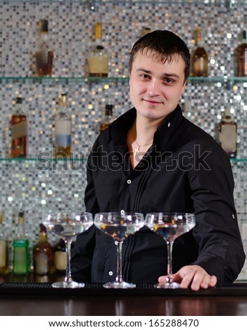 Smiling barman serving a batch of martini cocktails standing behind the bar counter smiling at the camera with the cocktails lined up in front of him