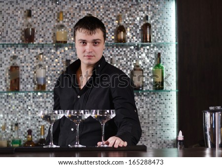 Three elegant saucer glasses with alcoholic beverages aligned on the bar by a professional Caucasian young bartender standing behing, in a fancy location