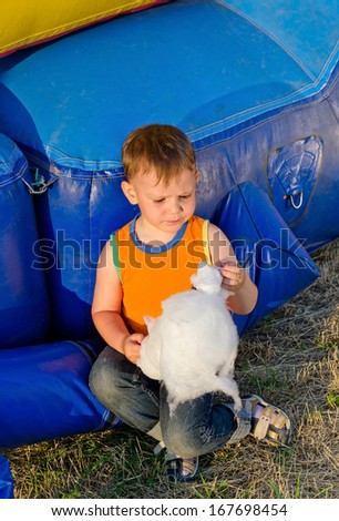 Small boy eating a portion of candy floss sitting on the ground in the sunshine alongside a colourful plastic jumping castle at a fairground