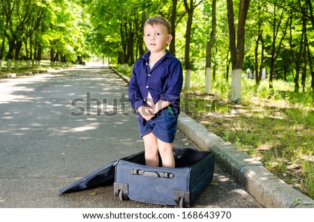 Cute young boy standing in an open suitcase in a rural street looking expectantly down the road as he waits for his lift to arrive to begin his summer vacation