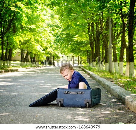 Young boy playing with an open suitcase on a rural road climbing inside as he waits in the shade of leafy green trees