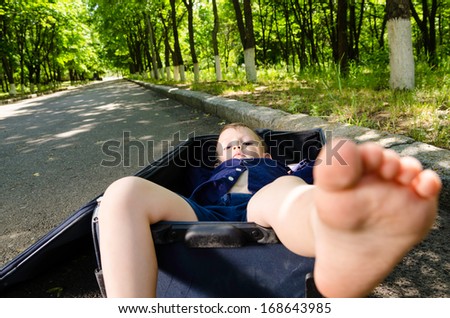 Barefoot little boy lying inside a suitcase on a rural road raising his head to look at the camera