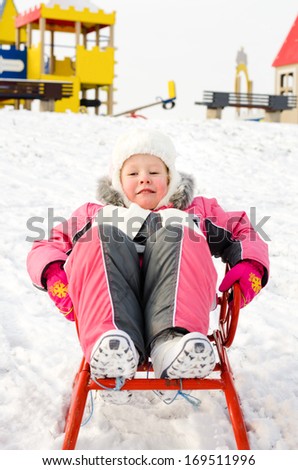 Pretty little girl dressed in a fashionable warm pink winter outfit playing on a sled in the snow reclining on her back smiling happily at the camera