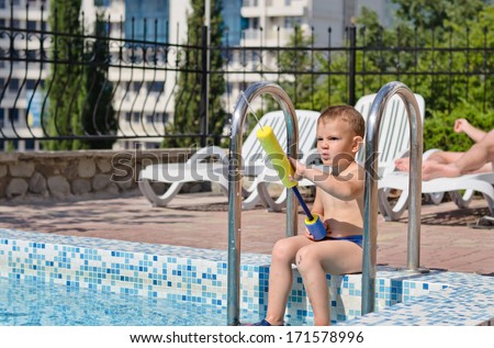Young boy enjoying the summer sun playing in a swimming pool sitting on the steps in his costume holding a colourful plastic toy with adults visible on loungers behind him
