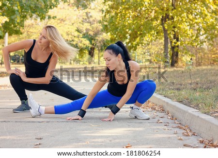 Two fit athletic young woman working out together on a road in a park doing stretching exercise and leg extensions to improve suppleness and mobility