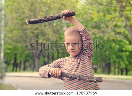 Cute little blond boy wearing camouflage pants standing playing with fighting sticks in a rural road practising his martial arts