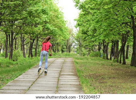 Athletic slender young teenage girl roller skating in a park on roller blades following a paved tree-lined path , view from behind