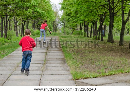 Little boy riding a scooter along a rural paved pathway following a teenage girl on roller blades who is skating ahead of him as the two enjoy some healthy exercise on a sunny day