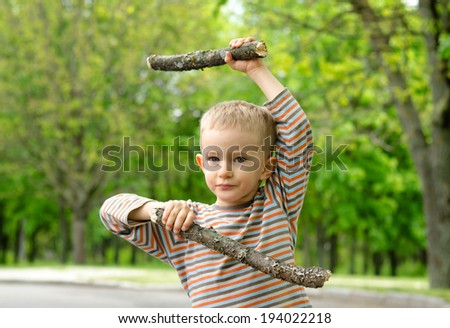 Cute little blond boy wearing camouflage pants standing playing with fighting sticks in a rural road practising his martial arts