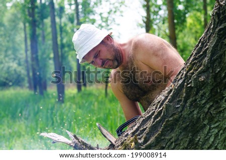 Fit shirtless man wearing a hat carrying a portable petrol chainsaw in woodland as he prepares to fell a tree for fuel