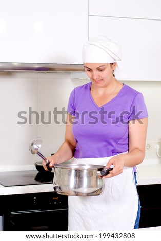 Female cook in a white apron and toque standing smelling her recipe in the pot which she has just removed from the hob