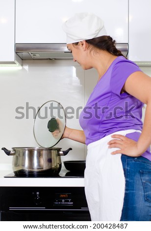 Female cook watching a pot boil on the stove in her white apron and chefs toque