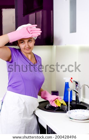 Tired or flustered female cook in a white apron and cap wiping her brow with her gloved hand as she does washing up at the sink