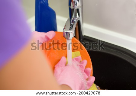 Woman rinsing plates under running water the tap as she does the washing up after a meal, close up of her gloved hands