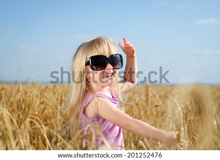 Adorable little blond girl with her hair tied up in pigtails posing in huge fashionable sunglasses in a golden wheat field laughing and waving