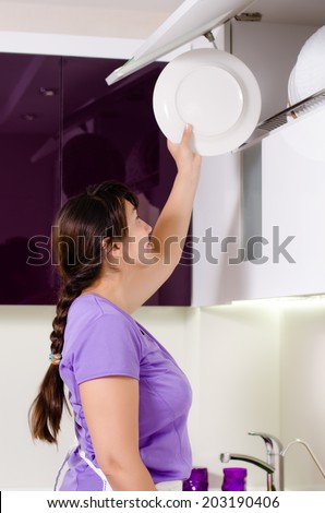 Attractive housewife doing the dishes standing at the kitchen sink reaching up to replace a dinner plate in storage in a rack in a cupboard