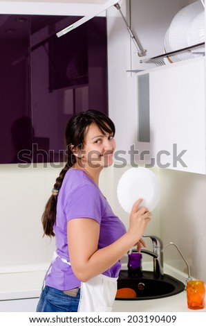 Attractive housewife doing the dishes standing at the kitchen sink reaching up to replace a dinner plate in storage in a rack in a cupboard