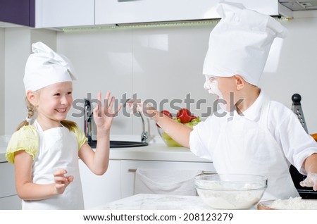 Happy little boy and girl baking in the kitchen in their cute white chefs uniforms laughing and fooling around with flour on their faces
