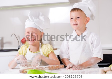 Happy little boy and girl wearing a white chefs uniform and hat cooking in the kitchen standing at the counter making a batch of biscuits and rolling the dough