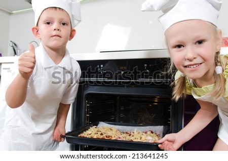 Happy excited young children dressed in white chefs uniforms grinning at the camera as they place a baking tray with homemade pizza in the oven that they have just made themselves