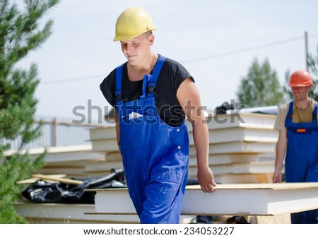Two young workmen carrying an insulated wooden wall panel on a building site as they move it from the stacked building supplies