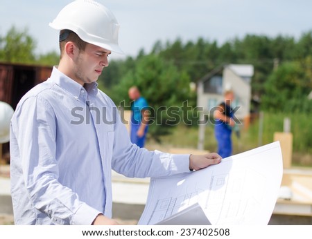 Architect or engineer checking plans on site holding the open blueprint in his hands as builders work in the background