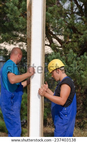 Two workmen in their hardhats and overalls installing insulation on a building site holding up a wooden insulated panel for the wall
