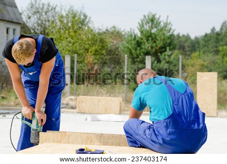 Two builders in dungarees installing insulated wall panels on a residential building site using an electric drill to drill holes in the wood