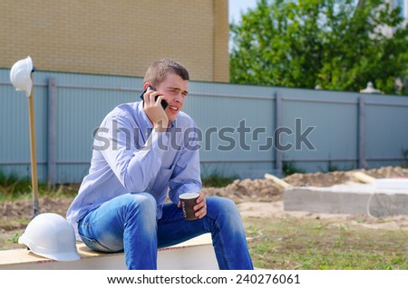 Young builder chatting on his mobile phone as he sits on building material on site enjoying a cup of coffee during a break