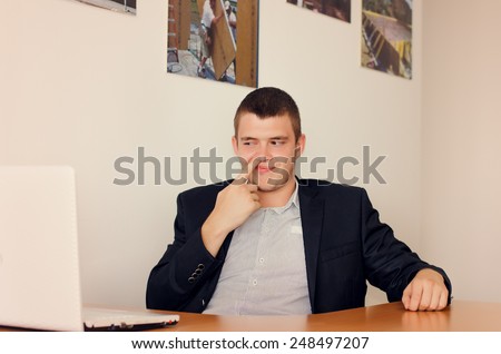 Young Businessman Sitting at Desk Picking Nose in Office