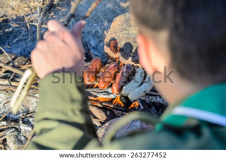 First Person View Looking Over Shoulder of Boy Scout Cooking Sausages on Stick over Campfire