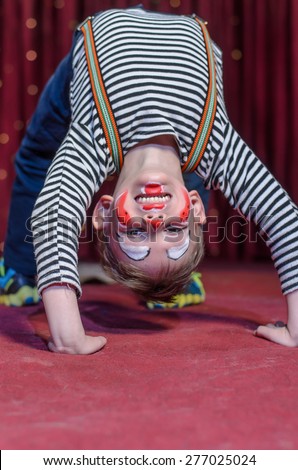 Supple agile little boy doing a back arch handstand on stage in a theatre wearing his colorful makeup as he grins at the camera from his upside down position
