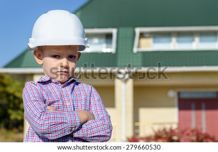 Young Boy Wearing Plaid Shirt and White Hard Hat Standing in front of House, Motioning with Hand as if Presenting Finished Home