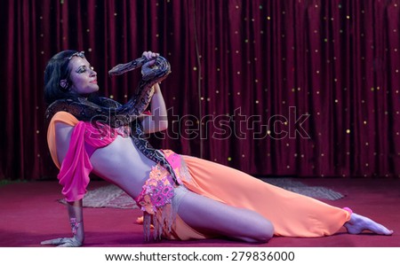 Full Length Image of Exotic Snake Charmer Dancer Reclining on Stage Floor with Large Snake Draped Around Shoulders, Fondly Looking at Snake Face to Face