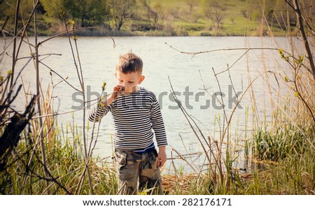 Young Boy Wearing Striped Shirt Stopping to Smell and Examine Plant Growing Amongst Reeds Along Shore of Lake