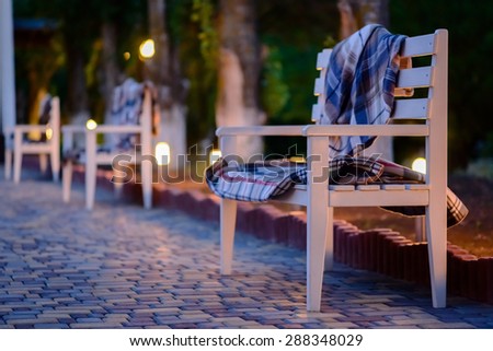 Blankets Piled on Empty Wooden Bench, Row of Benches on Stone Patio in Cozy Outdoor Setting Lit by Soft Evening Light
