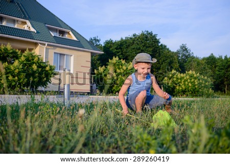Little boy catching insects with a net crouching down in the green grass outside the house with a look of anticipation on his face