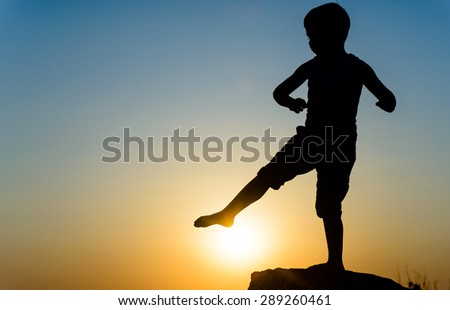 Little boy silhouetted on a wall or rock kicking the fiery orange orb of the setting sun with his foot, with copyspace