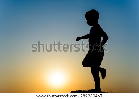 Little boy silhouetted on a wall or rock kicking the fiery orange orb of the setting sun with his foot, with copyspace