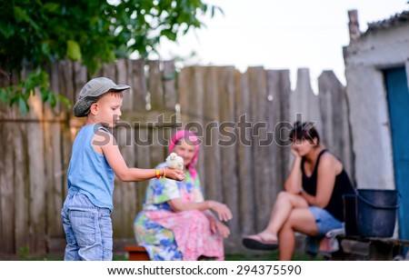 Young boy playing with a baby chicken holding it in the palm of his hand outdoors in the garden watched by his grandmother and mother