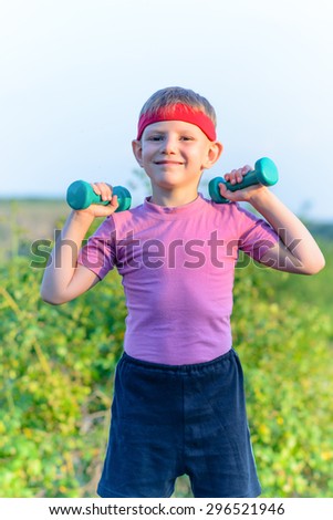 Close up Strong Young Boy with Warrior Headband, Lifting Two Small Dumbbells and Smiling at the Camera Against Greenery.