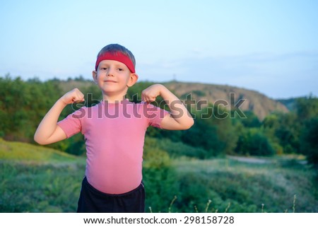 Little boy wearing a red headband standing outdoors showing off his biceps making a fist and flexing his arms