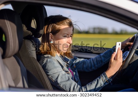 Young female driver reading a text message on her mobile phone as she drives along a rural road, distracting her attention from the road