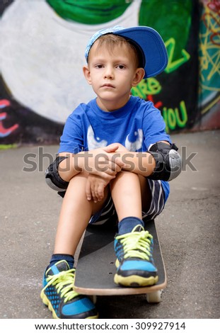 Portrait of Serious Young Boy Wearing Elbow Pads and Baseball Cap Sitting on Skateboard in Urban Setting with Graffiti Covered Wall in Background