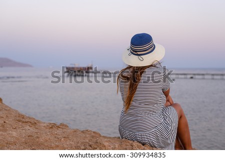 Romantic young woman wearing hat, striped dress and flip flops while sitting on the shore enjoying the breeze at the seaside in the morning