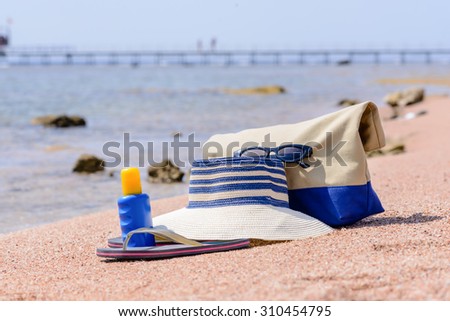 Ladies beach gear on a tropical beach with a sunhat, thongs, sunscreen and bag lying on golden sand overlooking the sea