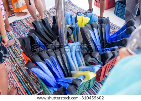 Box of different sized flippers or fins for hire by tourists at a resort with bare feet of diverse people standing in a queue visible